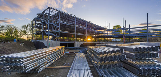 Steel beams and construction materials stacked in front of a partially built steel-framed building at sunset, with trees and sky in the background. Steel beams and construction materials stacked in front of a partially built steel-framed building at sunset, with trees and sky in the background.
